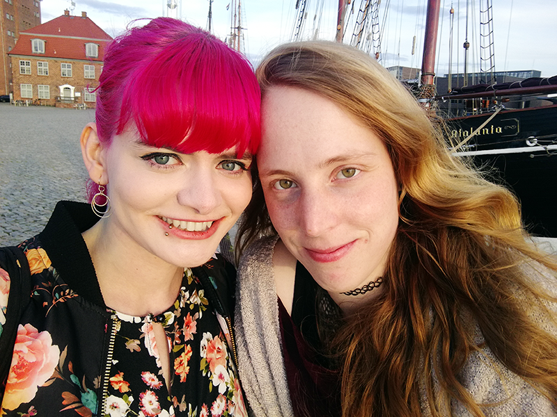 Lucia und ihre Partnerin Anna stehen am Wismarer Hafen und lächeln in die Kamera
