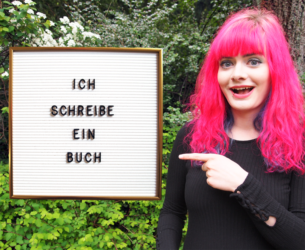Lucia steht draußen im Grünen und zeigt auf ein ins Bild gephotoshopptes Letterboard mit der Aufschrift: Ich schreibe ein Buch.