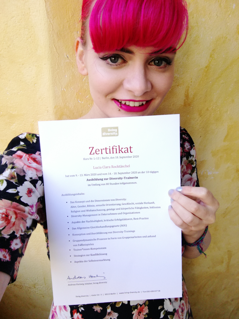 Lucia hält ihr Zertifikat der Ausbildung zur Diversity-Trainerin in der Hand und lächelt in die Kamera.