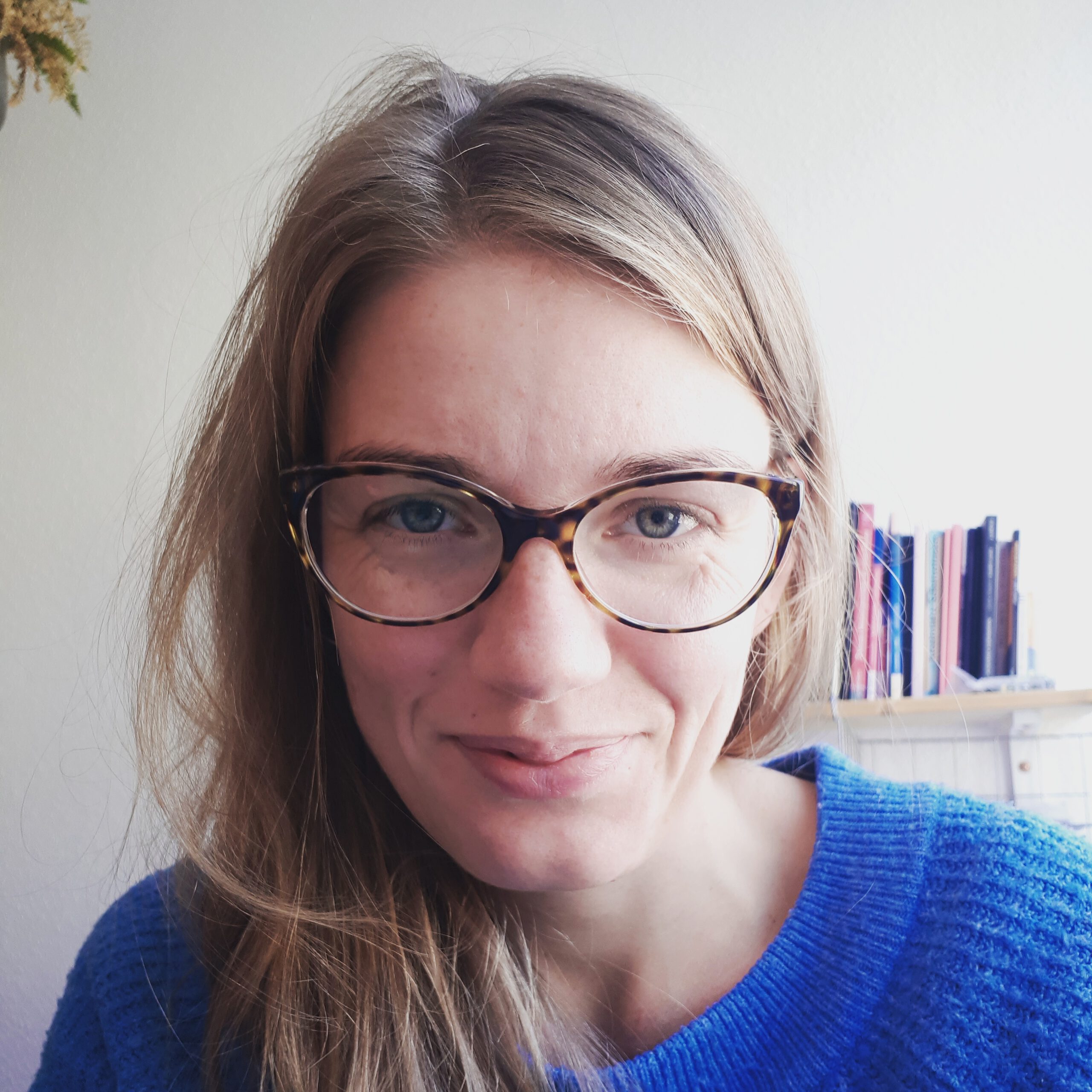 Porträt von Nora Stankewitz. Sie hat lange naturblonde Haare, trägt eine Brille und lächelt in die Kamera. Im Hintergrund ist ein Bücherregal zu sehen.