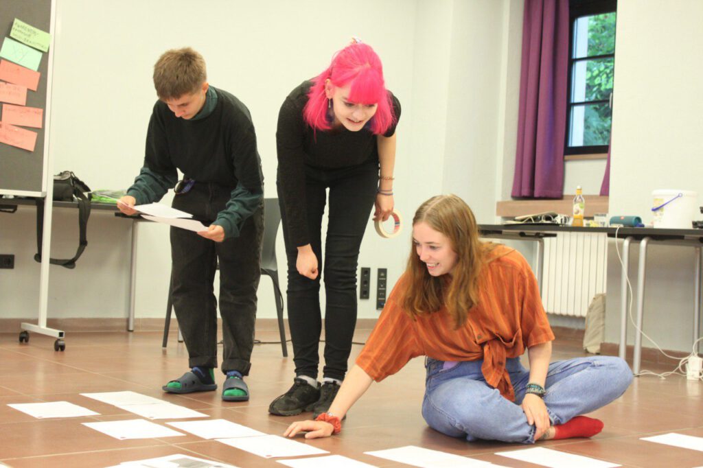 Das Careleaving-Storys-Team aus Lee, Lucia und Laura beugt sich über lose Blätter, die auf dem Boden liegen. Sie bringen die Blätter mit den Geschichten aus der Jugendhilfe in die richtige Reihenfolge.